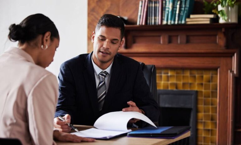 A lawyer sits at his desk and explains a legal document to a client while he points at it with a pen.